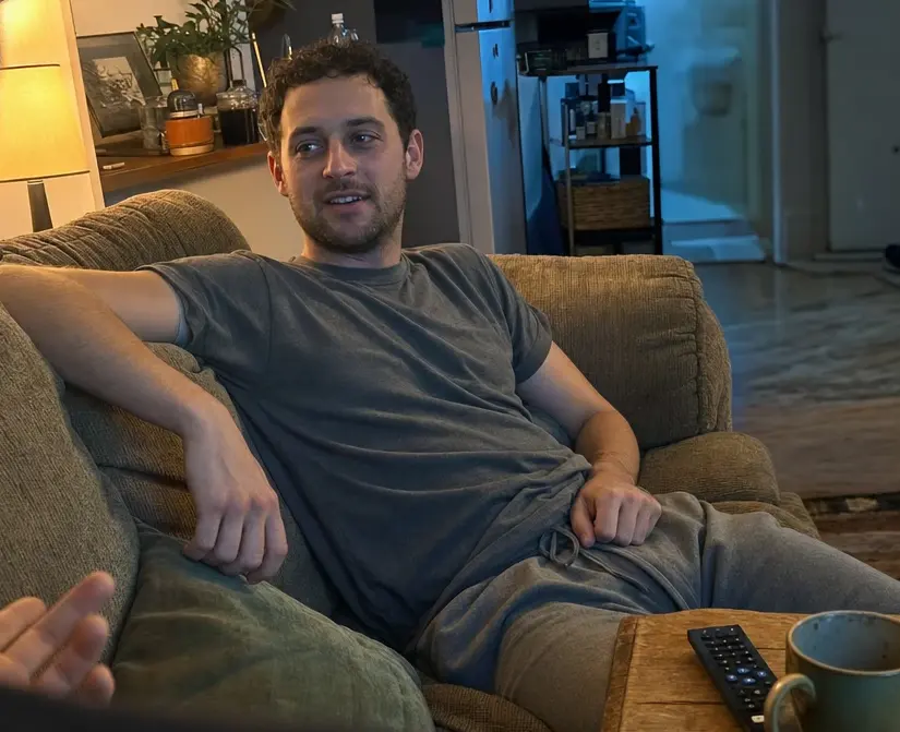 A man sunk into a worn couch at home in the evening, relaxed and mid-conversation, unaware he is being photographed