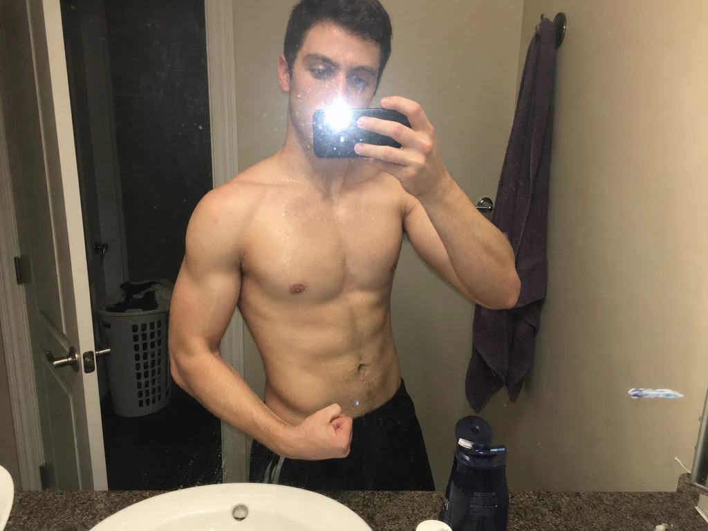 A man taking a shirtless bathroom mirror selfie at night with a harsh camera flash, posed in a stiff flex