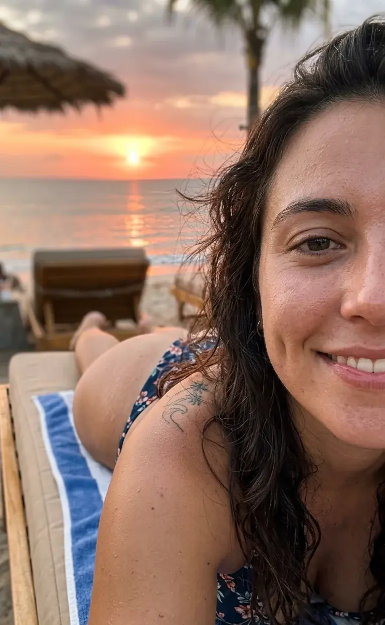A smiling woman takes a sunset selfie lying on a beach lounger, her bikini-clad figure visible in the background of the frame